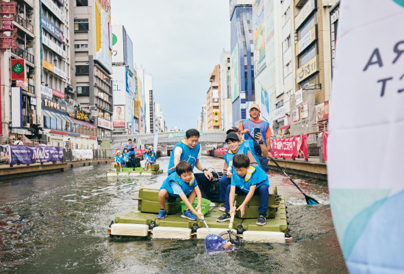 道頓堀川で、いかだのようなグループで乗れるサップボートに乗った親子が網を使って浮遊ごみを回収している様子