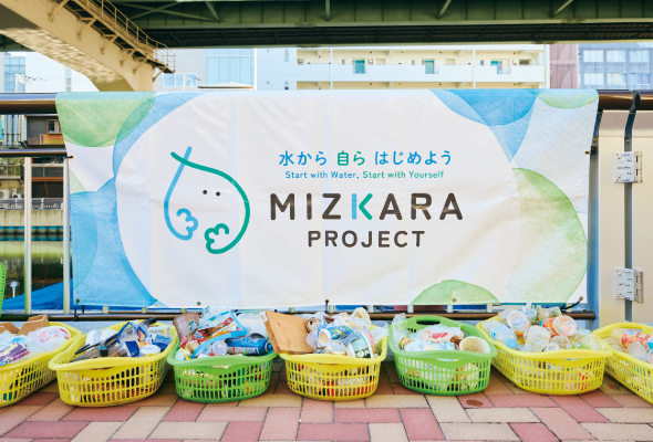 ミズカラプロジェクトのロゴが印刷された横断幕の前に、水辺キレイツアーで拾ったごみをカゴに入れて集めた様子